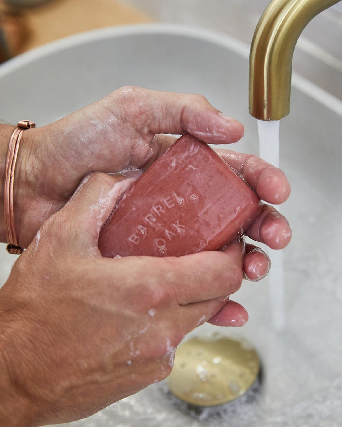 Hands lathering a Barrel and Oak Bourbon Cedar exfoliating soap bar under running water from a gold faucet.