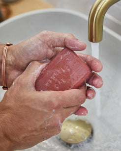 Hands lathering a Barrel and Oak Bourbon Cedar exfoliating soap bar under running water from a gold faucet.