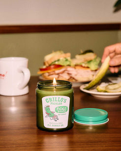 P.F. Candle Co. Grillo’s Pickle Scented Candle lit on a table next to a sandwich and coffee mug – limited edition green soy candle inspired by Grillo’s Pickles