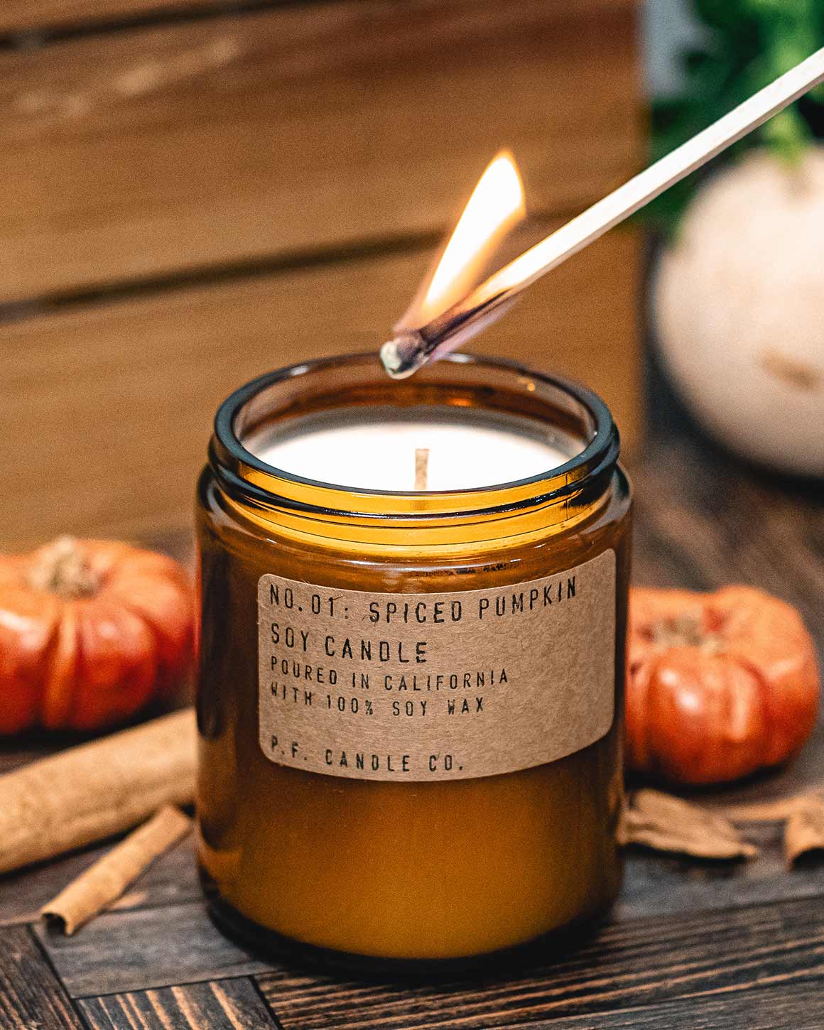 Spiced pumpkin candle being lit with a match, surrounded by pumpkins and cinnamon sticks on a wooden surface.