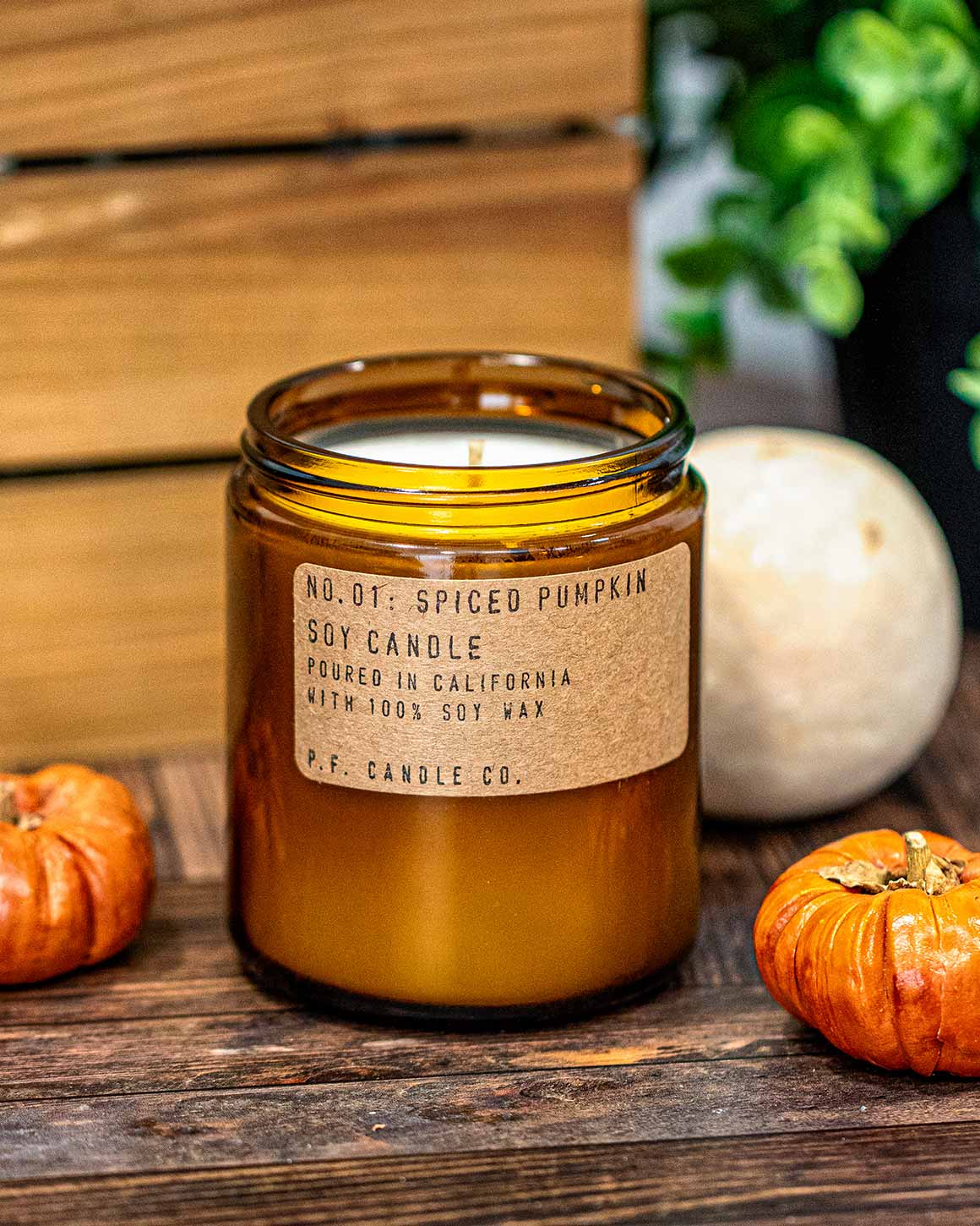 Spiced pumpkin candle in a amber jar with decorative pumpkins and apples on a wooden surface.