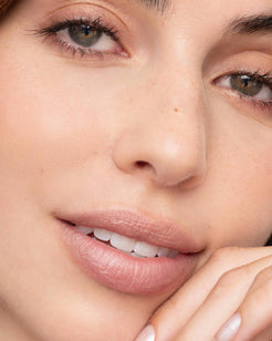 Close-up of a woman's face showing natural, matte pink lipstick on her lips, with clear skin and subtle freckles.